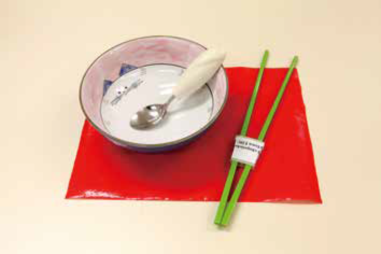 A red non-slip mat. On the mat, there is a spoon with build- up handle in the bowl, by the side is chopsticks with chopsticks adaptor. 