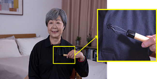 A woman buttoning a shirt using buttoning aid. The buttoning aid has a handle, and a metal loop at the tip. The buttoning aid would be inserted into the button hole of the shirt, and the button would be hooked by the metal loop, and finally the aid would be pulled out from the hole of the shirt.